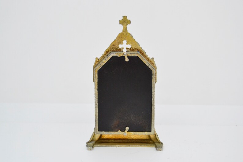 May include: Ornate gold-colored metal picture frame with a cross at the top. The frame has a dark, rectangular insert and a decorative, architectural design. The frame is standing upright on a white surface.