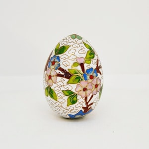 May include: A white egg-shaped decorative object with a floral design. The design features pink, blue, and red flowers with green leaves and gold accents.