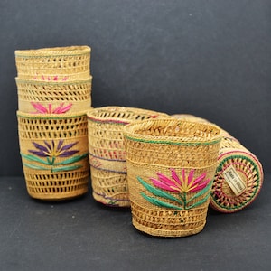 Puede incluir: Conjunto de cinco cestas tejidas con diseños florales en rosa, verde y morado. Las cestas están hechas de materiales naturales y tienen un aspecto rústico. La etiqueta en una cesta dice "Hecho en China."
