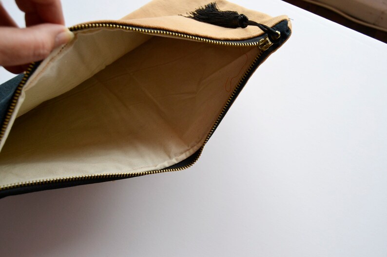 May include: A tan and black zippered pouch with a black tassel. The pouch is open and the inside is lined with a cream-colored fabric.