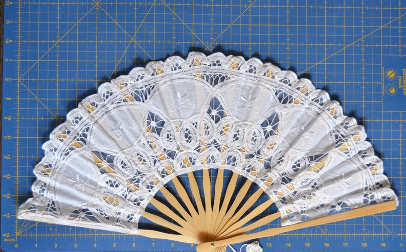 May include: A white lace fan with a wooden handle. The fan is closed and the lace is intricate and delicate.