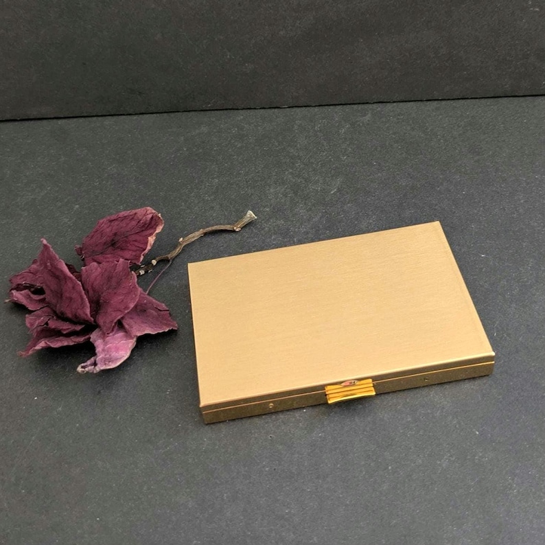Puede incluir: Un estuche compacto rectangular de color dorado con un acabado de metal cepillado. El estuche tiene un peque&ntilde;o cierre dorado. Una flor seca de color p&uacute;rpura oscuro y una ramita peque&ntilde;a est&aacute;n junto al compacto.