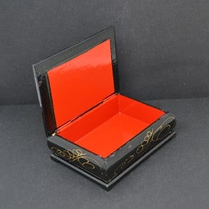 May include: A black wooden box with a red interior and gold painted floral designs. The box is open and the lid is propped up.