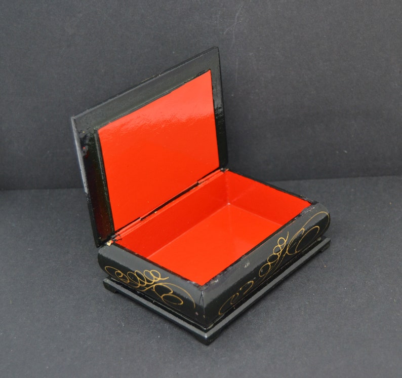 May include: A black wooden box with a red interior and gold painted floral designs. The box is open and the lid is propped up.