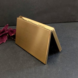 Puede incluir: Un tarjetero rectangular de metal dorado. El soporte est&aacute; abierto, con un lado inclinado hacia arriba, revelando un interior oscuro. La superficie tiene una textura cepillada y los bordes son limpios y afilados.