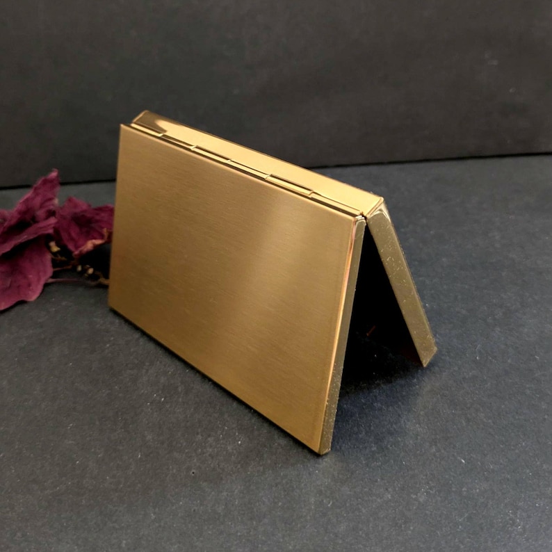 Puede incluir: Un tarjetero rectangular de metal dorado. El soporte est&aacute; abierto, con un lado inclinado hacia arriba, revelando un interior oscuro. La superficie tiene una textura cepillada y los bordes son limpios y afilados.
