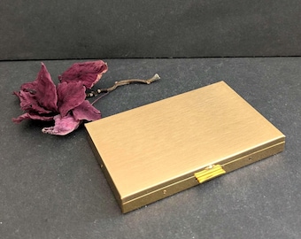 May include: A rectangular, gold-colored metal case with a brushed finish. The case has a small, gold-colored clasp and is next to a dried, dark red flower and a small twig. The background is dark gray.