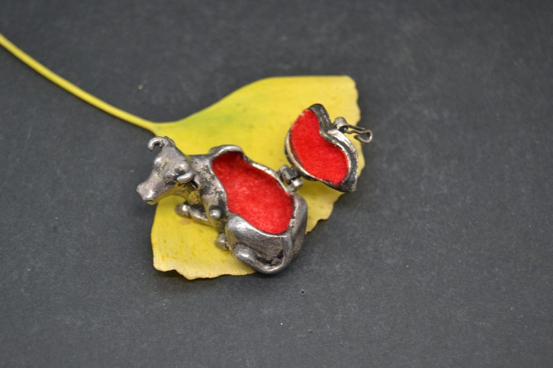 May include: A silver cow-shaped trinket box with a red velvet interior. The box is open and the lid is propped up on a yellow leaf.