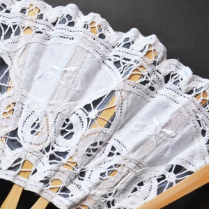 May include: A white lace fan with a wooden handle. The fan is decorated with a delicate lace pattern and has a white fabric center.