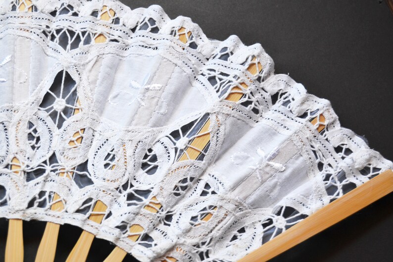 May include: A white lace fan with a wooden handle. The fan is decorated with a delicate lace pattern and has a white fabric center.