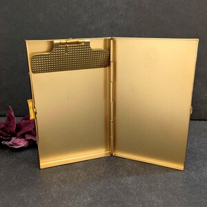 Puede incluir: Estuche met&aacute;lico dorado abierto, con un clip y una bisagra texturizados. La caja es rectangular y tiene una superficie lisa y reflectante. El interior tambi&eacute;n es dorado, con un dise&ntilde;o simple y elegante.