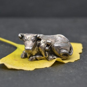 May include: A silver cow figurine resting on a yellow leaf. The cow is lying down with its head turned to the side.