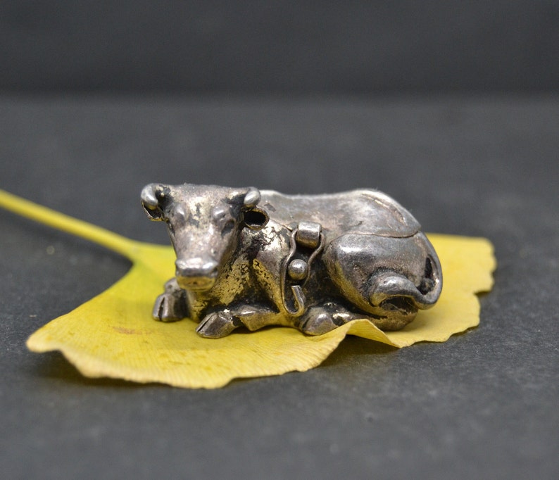 May include: A silver cow figurine resting on a yellow leaf. The cow is lying down with its head turned to the side.