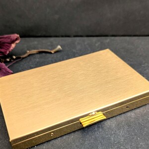 Puede incluir: Un estuche rectangular de metal dorado con acabado cepillado. El estuche tiene un peque&ntilde;o cierre dorado y est&aacute; cerrado. El fondo es una superficie gris oscuro con unos p&eacute;talos de flores secas.