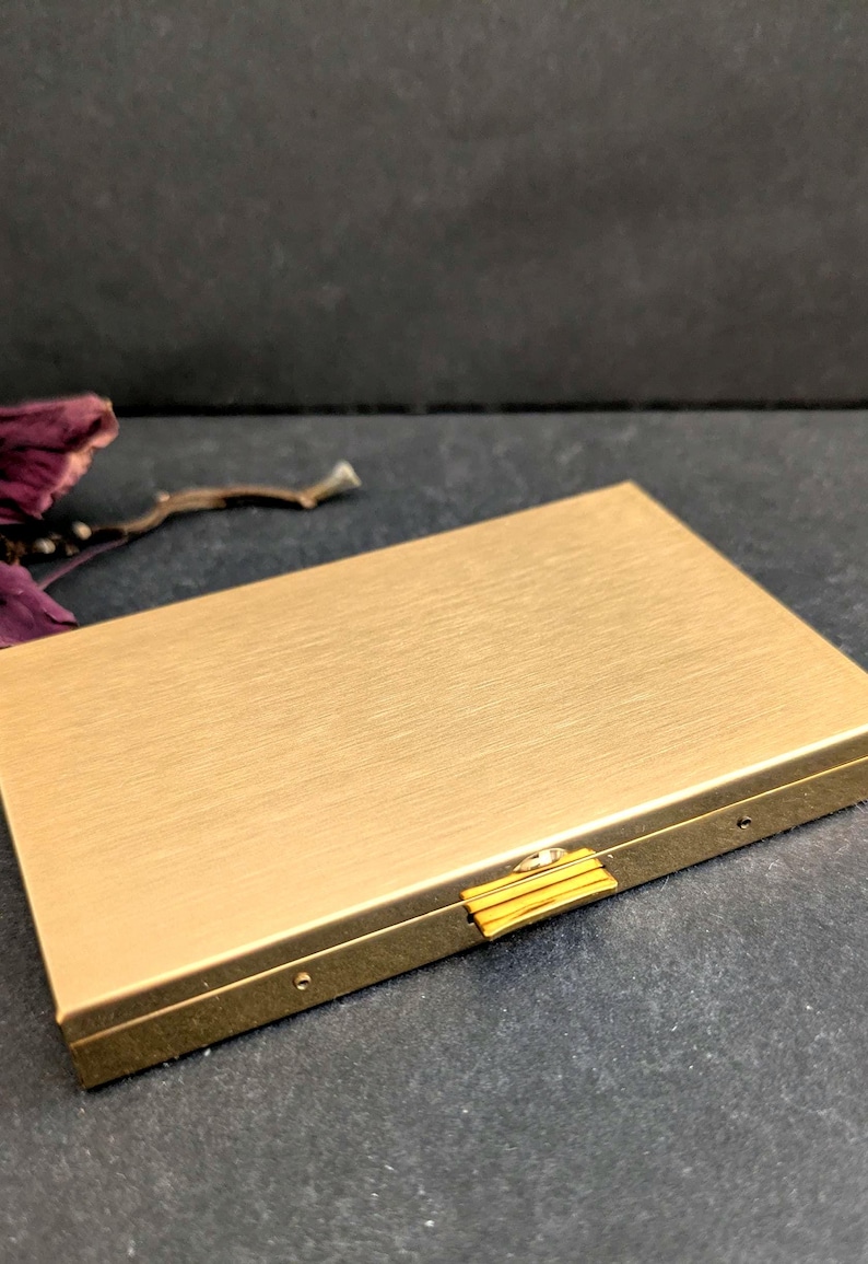 Puede incluir: Un estuche rectangular de metal dorado con acabado cepillado. El estuche tiene un peque&ntilde;o cierre dorado y est&aacute; cerrado. El fondo es una superficie gris oscuro con unos p&eacute;talos de flores secas.