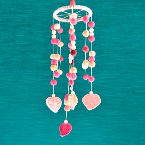 May include: A pink and white baby mobile with crocheted hearts and pom-poms hanging from a wooden ring.