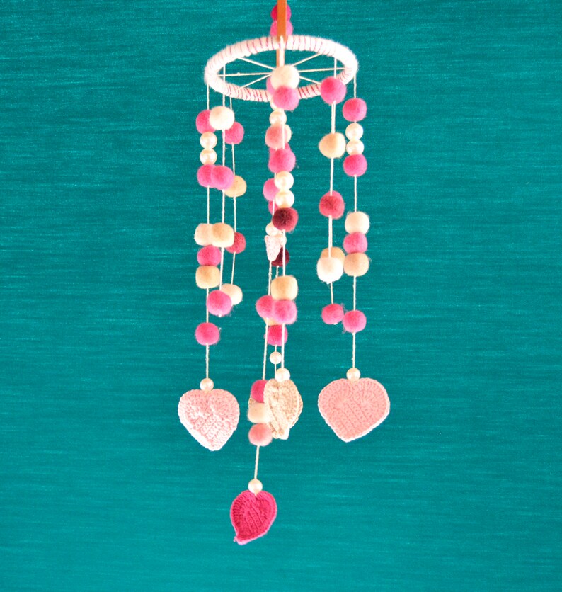 May include: A pink and white baby mobile with crocheted hearts and pom-poms hanging from a wooden ring.