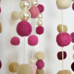 May include: A pink and cream felt ball mobile with white pearl beads hanging from a white ceiling. The mobile is made of felt balls in various sizes and colors, including pink, cream, and white.