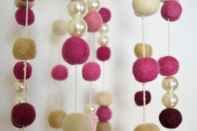 May include: A pink and cream felt ball mobile with white pearl beads hanging from a white ceiling. The mobile is made of felt balls in various sizes and colors, including pink, cream, and white.