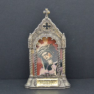 May include: A silver-toned religious icon featuring an image of Jesus wearing a crown of thorns, set against a red and gold background. The icon has an ornate, arched frame with a cross at the top and a small plaque with text at the bottom.