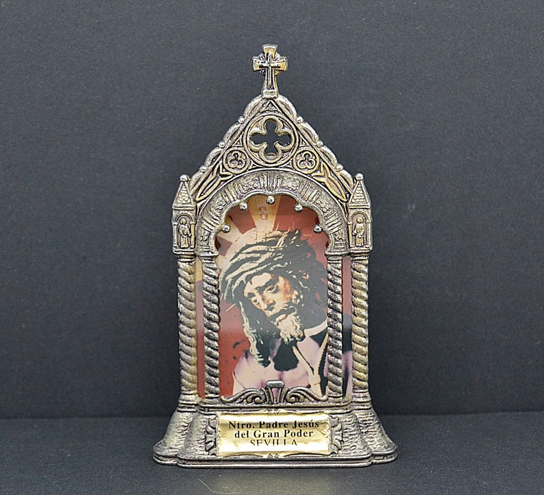May include: A silver-toned religious icon featuring an image of Jesus wearing a crown of thorns, set against a red and gold background. The icon has an ornate, arched frame with a cross at the top and a small plaque with text at the bottom.