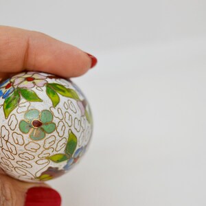 May include: A white cloisonn&eacute; ball with a floral design in shades of green, blue, pink, and red. The ball is decorated with a repeating pattern of gold lines.