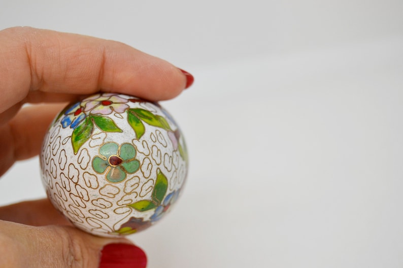 May include: A white cloisonn&eacute; ball with a floral design in shades of green, blue, pink, and red. The ball is decorated with a repeating pattern of gold lines.