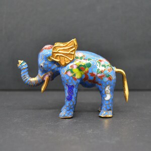 Puede incluir: Una pequeña figurita de elefante de cloisonné azul y dorado con intrincados diseños florales. El elefante tiene la trompa levantada y una cola dorada.