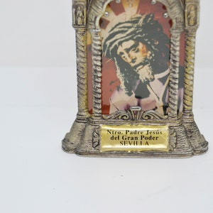 May include: A silver-toned religious icon featuring an image of Jesus with a crown of thorns. The icon has a decorative frame with a gold-colored plaque at the base that reads "Ntro. Padre Jes&uacute;s del Gran Poder SEVILLA."