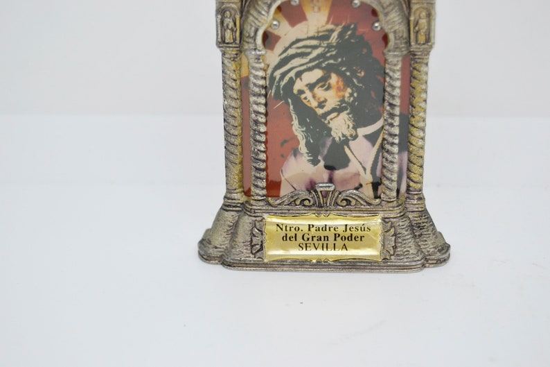 May include: A silver-toned religious icon featuring an image of Jesus with a crown of thorns. The icon has a decorative frame with a gold-colored plaque at the base that reads "Ntro. Padre Jes&uacute;s del Gran Poder SEVILLA."