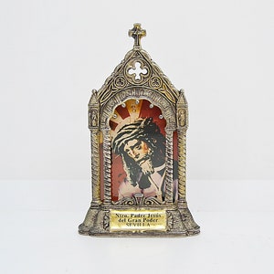 May include: A religious icon in a decorative metal frame. The frame is silver and gold-toned, with a cross at the top. The image depicts a portrait of Jesus with a crown of thorns, set against a red and gold background. The text "Ntro. Padre Jes&uacute;s del Gran Poder SEVILLA" is at the bottom.