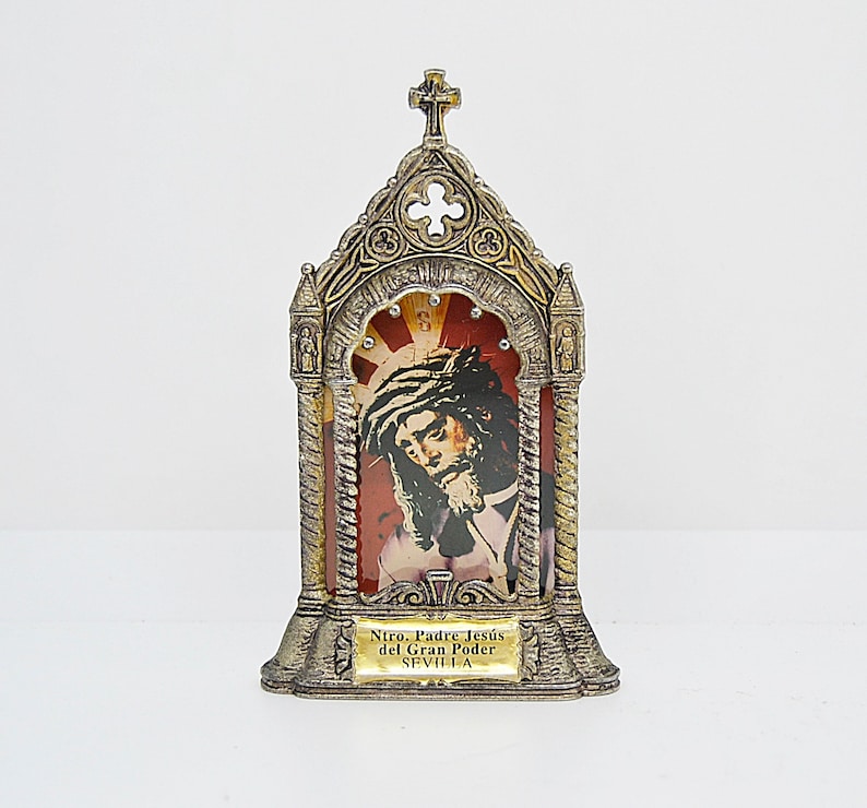 May include: A religious icon in a decorative metal frame. The frame is silver and gold-toned, with a cross at the top. The image depicts a portrait of Jesus with a crown of thorns, set against a red and gold background. The text "Ntro. Padre Jes&uacute;s del Gran Poder SEVILLA" is at the bottom.
