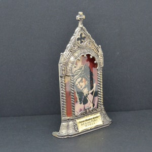 May include: A silver-colored religious icon featuring an image of a man with a beard and a crown of thorns. The icon has a decorative frame with a cross at the top and the text "Ntro. Padre Jes&uacute;s del Gran Poder SEVILLA" on a small plaque.