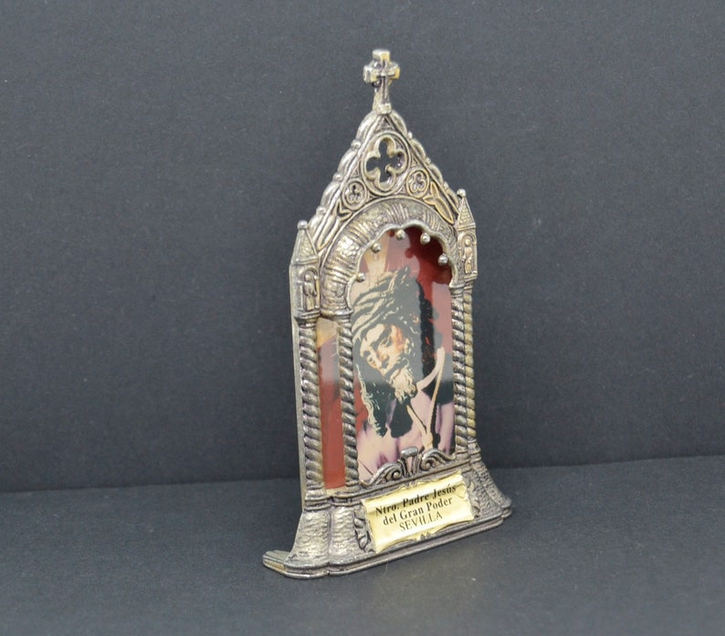 May include: A silver-colored religious icon featuring an image of a man with a beard and a crown of thorns. The icon has a decorative frame with a cross at the top and the text "Ntro. Padre Jes&uacute;s del Gran Poder SEVILLA" on a small plaque.