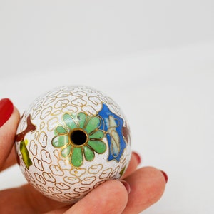 May include: A white cloisonn&eacute; ball with a hole in the center. The ball is decorated with a green, blue, and gold floral design.
