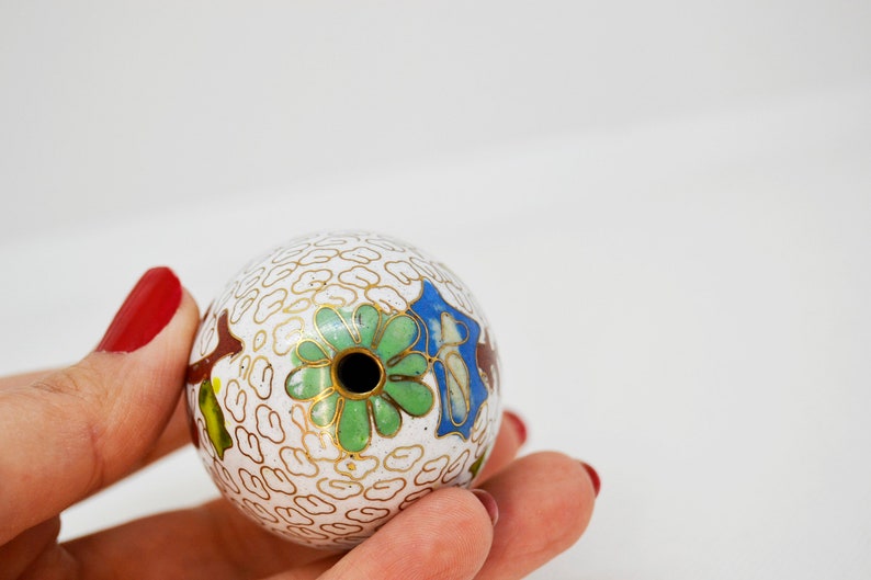 May include: A white cloisonn&eacute; ball with a hole in the center. The ball is decorated with a green, blue, and gold floral design.