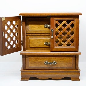 Puede incluir: Una caja de madera para joyas con una puerta con bisagras y tres cajones. La caja tiene un diseño de celosía en la puerta y los cajones, y asas de metal. La caja es marrón y tiene un acabado desgastado.