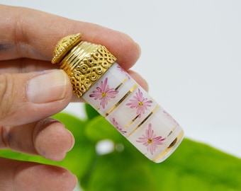 Op de afbeelding: Een klein, wit glazen flesje met een gouden dop en een roze bloemenpatroon. Het flesje heeft gouden strepen en is versierd met roze bloemen.