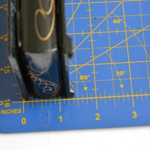 May include: A black and gold book with a decorative design on the cover. The book is measured on a blue cutting mat with a ruler marked in inches.