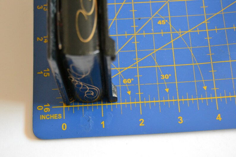 May include: A black and gold book with a decorative design on the cover. The book is measured on a blue cutting mat with a ruler marked in inches.