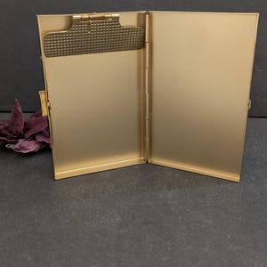 Puede incluir: Portatarjetas de visita de metal dorado abierto. El soporte es rectangular con un clip texturizado en el lado izquierdo. El interior es de color dorado liso. El soporte est&aacute; abierto, revelando el interior.