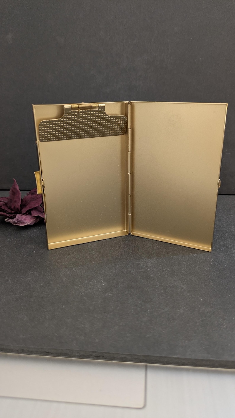 Puede incluir: Portatarjetas de visita de metal dorado abierto. El soporte es rectangular con un clip texturizado en el lado izquierdo. El interior es de color dorado liso. El soporte est&aacute; abierto, revelando el interior.