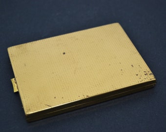 May include: A gold-colored metal compact with a textured surface. The compact has a small latch on the side.