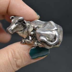 May include: A silver cow figurine with a detailed design. The cow is lying down with its head turned to the side. The figurine is about 2 inches long.