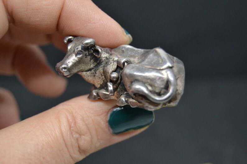 May include: A silver cow figurine with a detailed design. The cow is lying down with its head turned to the side. The figurine is about 2 inches long.
