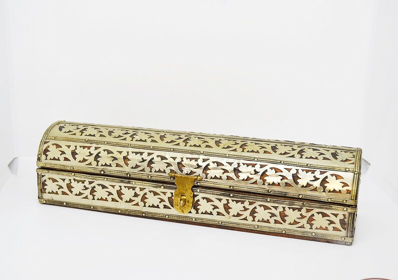 May include: A long, rectangular box with a curved top. The box is made of wood and has intricate silver and gold floral designs carved into the surface. The box has a brass latch.