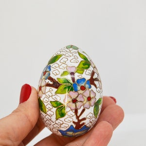 May include: A white egg-shaped trinket box with a floral design in shades of green, blue, pink, and gold. The box is decorated with a delicate pattern of flowers and leaves.