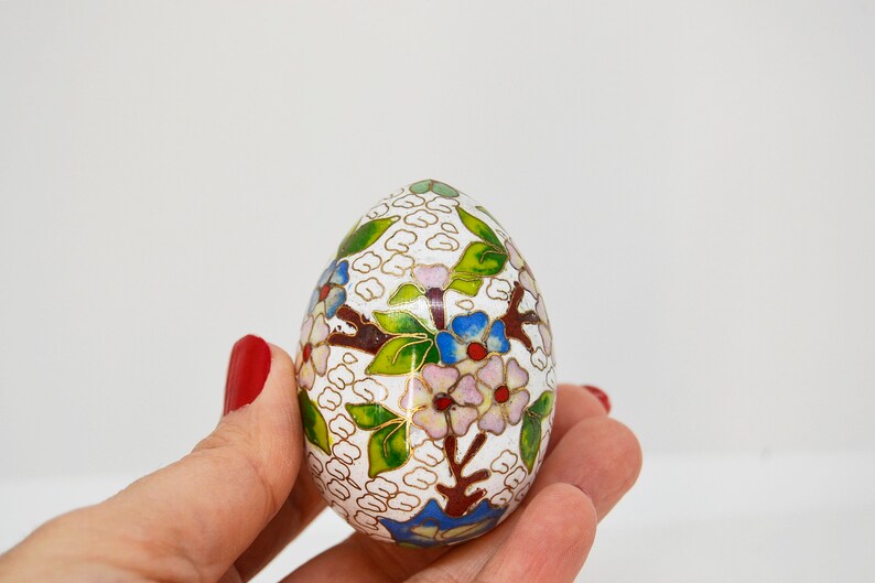 May include: A white egg-shaped trinket box with a floral design in shades of green, blue, pink, and gold. The box is decorated with a delicate pattern of flowers and leaves.