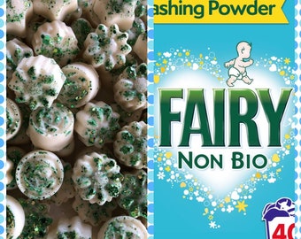 May include: A close-up of a pile of white wax melts with green glitter. The melts are shaped like stars, flowers, and swirls. The image is next to a box of Fairy Non Bio washing powder. The box is blue with a white heart and the words "Fairy Non Bio" in green letters.