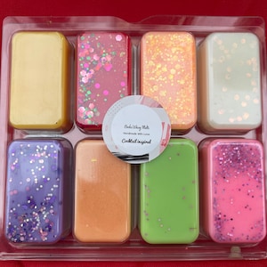 May include: Eight rectangular wax melts in a clear plastic packaging. The wax melts are in various colours and have glitter embedded in them. The packaging has a label that reads "Books & Wax Melts Handmade with Love Cocktail Inspired".